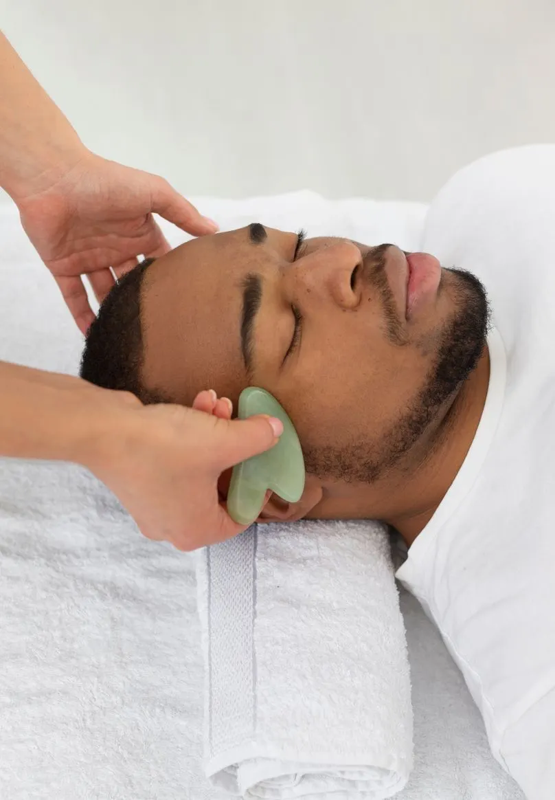 A therapist works a jade gua sha across a client's cheek during a facial.